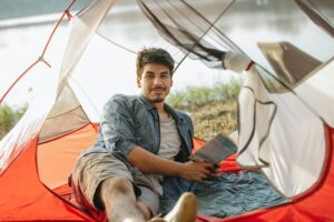 cheerful man reading book in camp tent near calm lake